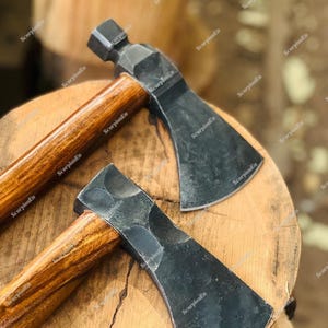Könnte beinhalten: Zwei handgefertigte Äxte mit dunklen Metallköpfen und Holzgriffen. Die Äxte werden auf einer Holzoberfläche präsentiert, die ihr rustikales Design hervorhebt. Die Griffe haben eine satte, braune Oberfläche, und die Axtköpfe haben ein strukturiertes, dunkles Aussehen.