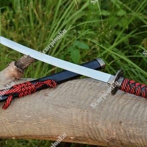 Könnte beinhalten: Ein Katana-Schwert mit silberner Klinge und einem rot-schwarzen Griff. Das Schwert liegt auf einem Holzstamm, daneben die Scheide. Die Scheide ist schwarz mit roten Akzenten.