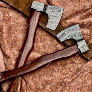 May include: Two axes with patterned steel blades and dark wooden handles rest on a brown surface. The blades have a unique swirling pattern, and the handles are a rich, dark brown. The axes appear to be handcrafted.