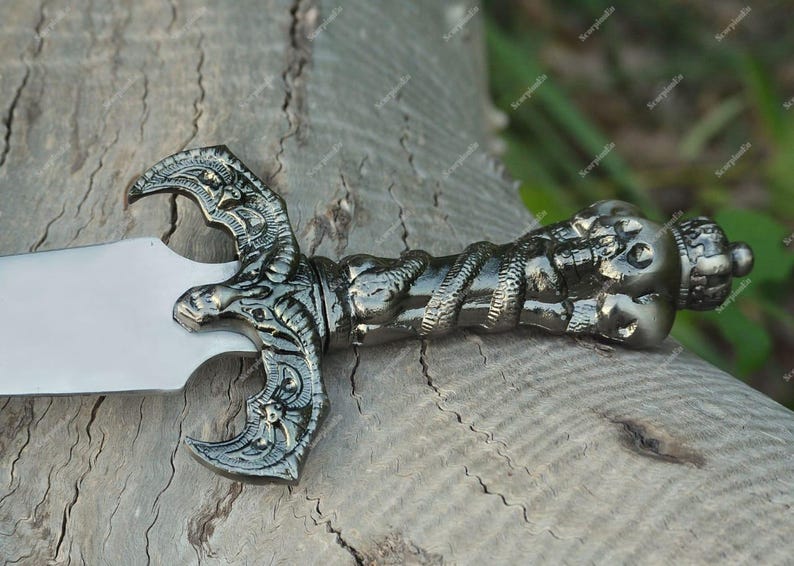 May include: A detailed, silver-colored sword with an ornate handle and a wide, polished blade. The handle features intricate carvings and a decorative pommel. The sword is resting on a weathered wooden surface, showcasing its craftsmanship.
