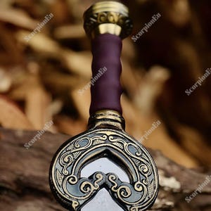 Può includere: Primo piano di una spada decorativa. La spada presenta un'impugnatura viola e una guardia dorata con intricati motivi. La lama è trasparente, forse di vetro. Il design suggerisce un tema storico o fantasy, con attenzione ai dettagli.