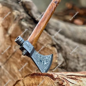 May include: A handcrafted tomahawk axe with a patterned steel blade and a wooden handle. The handle is a rich brown color, and the blade has a unique swirling design. The axe is resting on a piece of wood.