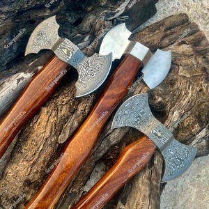 Könnte beinhalten: Vier Äxte mit gemusterten Stahlklingen und Holzgriffen. Die Klingen haben ein einzigartiges Design und die Griffe sind in einem satten, dunklen Braun gehalten. Die Äxte sind auf einer strukturierten Holzoberfläche angeordnet.