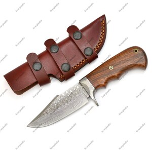 May include: A hunting knife with a patterned blade and a wooden handle, accompanied by a brown leather sheath. The sheath has three straps with metal studs. The knife blade is approximately 5 inches long.