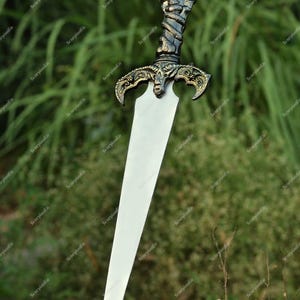 May include: A decorative sword with a silver blade and an ornate handle. The handle features a skull and serpent design, with gold-colored accents. The sword is displayed against a blurred green background.