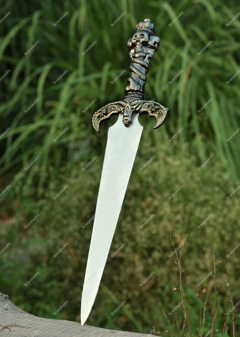 May include: A decorative sword with a silver blade and an ornate handle. The handle features a skull and serpent design, with gold-colored accents. The sword is displayed against a blurred green background.
