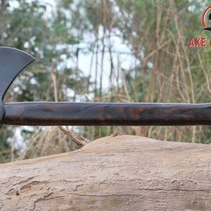 Pode incluir: Machado com cabo de madeira escura e cabe&ccedil;a preta. A cabe&ccedil;a do machado tem uma l&acirc;mina afiada e uma ponta no lado oposto. O cabo &eacute; curvo para a ader&ecirc;ncia. O machado est&aacute; apoiado em um tronco.