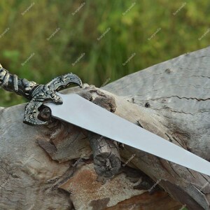 May include: A decorative dagger with a silver blade and an ornate handle. The handle features a detailed design with a skull and other decorative elements. The dagger is resting on a weathered wooden log.