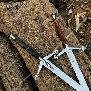 Könnte beinhalten: Zwei lange Schwerter mit silbernen Klingen und verzierten Parierstangen liegen auf einer strukturierten Holzoberfläche. Ein Schwert hat einen schwarz umwickelten Griff, das andere einen braunen, gedrehten Griff. Die Knäufe sind silberfarben und die Klingen poliert.