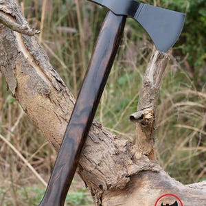 Pode incluir: Machado com cabo de madeira escura e polida e l&acirc;mina preta. O machado est&aacute; apoiado em um galho de &aacute;rvore desgastado, com o logotipo Axe Master vis&iacute;vel no canto inferior direito. O cabo tem uma extremidade curva para uma pegada segura.
