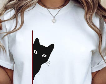 May include: White t-shirt featuring a black cat graphic peeking from behind a red line. The cat has white eyes and whiskers. The shirt is a casual style.