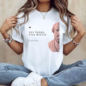 May include: White t-shirt featuring the quote "Ars longa, Vita Brevis" and a bust of Hippocrates. The design is in black and a muted pink. The shirt is a classic crew neck style.