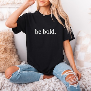 May include: Black t-shirt with the words "be bold." in white, featuring a crew neck and short sleeves. The shirt is paired with light blue ripped jeans. The overall style is casual and comfortable.