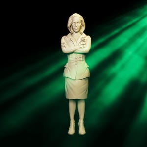 Op de afbeelding: Gedetailleerd beeldje van een vrouw in een lichtgekleurd pak met gekruiste armen. Het beeldje heeft een rok, een blazer en een kleine badge met de tekst "FBI". De achtergrond is donker met groene lichtstralen.