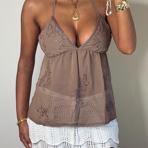May include: A brown halter top with floral embroidery and a white crochet skirt. The top has a plunging neckline and a flowing, sheer fabric. The skirt has a scalloped hemline. Accessories include a gold necklace and bracelets.