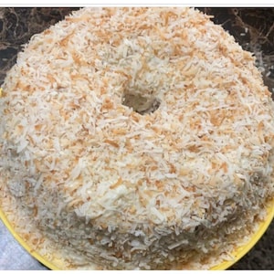 May include: A round coconut cake with a hole in the center, covered in shredded coconut. The cake sits on a yellow plate, with a dark brown and black marble-like background.