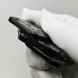 以下が含まれることがあります： 金と銀のアクセントが付いた黒い金属製のフィジェットスピナー。スピナーの表面には詳細なデザインが施されています。画像は側面から撮影されており、積層構造と複雑なディテールが示されています。