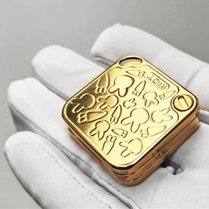 以下が含まれることがあります： 金色の正方形のコンパクト。ヒンジ付きの蓋には、歯、ニンジン、キノコの漫画のエンボス加工が施されています。「MOT」の文字が見えます。手袋をはめた手でコンパクトが持たれています。
