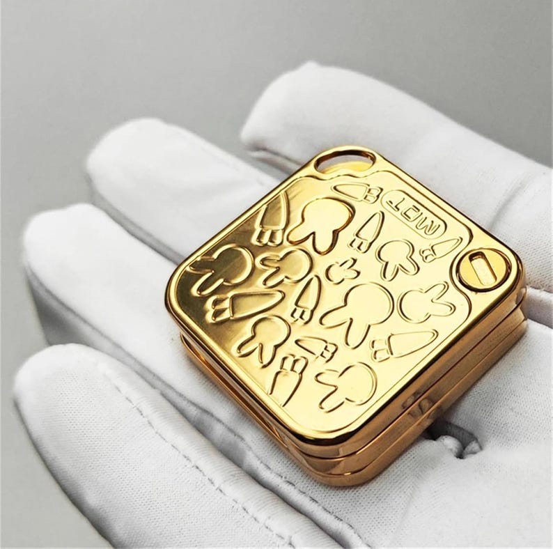 以下が含まれることがあります： 金色の正方形のコンパクト。ヒンジ付きの蓋には、歯、ニンジン、キノコの漫画のエンボス加工が施されています。「MOT」の文字が見えます。手袋をはめた手でコンパクトが持たれています。