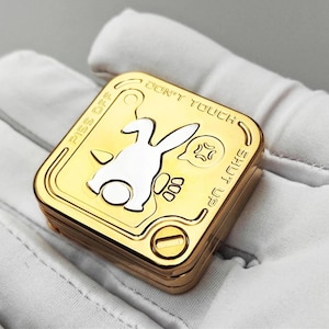 以下が含まれることがあります： 金色の正方形の小物入れ。白い漫画のウサギのデザイン。ボックスには「PISS OFF」、「DON'T TOUCH」、「SHUT UP」のフレーズが刻まれています。ウサギは逆さまになっていて、怒った顔をしています。