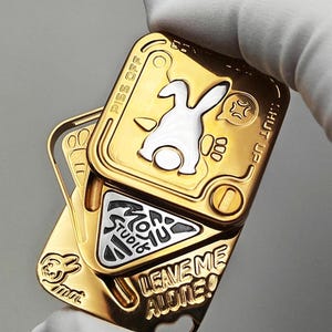 以下が含まれることがあります： ヒンジデザインの金色の金属製フィジェットおもちゃ。おもちゃには白いウサギのグラフィック、「PISS OFF」、「HUT UP」、および「LEAVE ME ALONE！」のテキストが特徴です。中央には「MON YU STUDIO」というテキストが入った三角形があります。