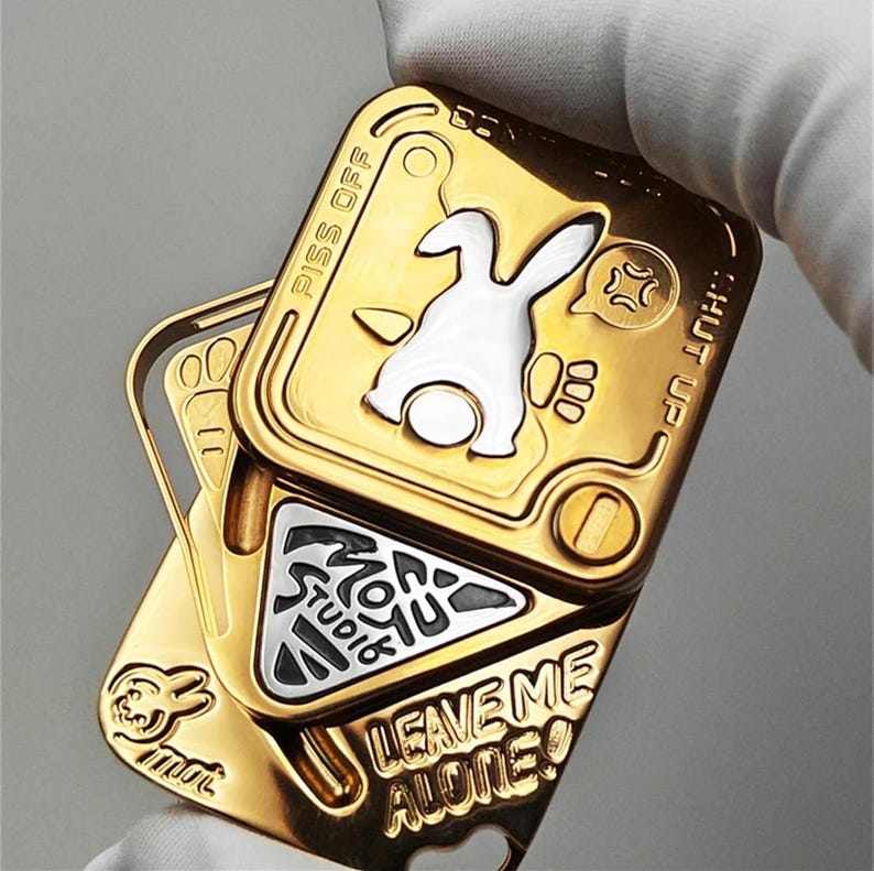 以下が含まれることがあります： ヒンジデザインの金色の金属製フィジェットおもちゃ。おもちゃには白いウサギのグラフィック、「PISS OFF」、「HUT UP」、および「LEAVE ME ALONE！」のテキストが特徴です。中央には「MON YU STUDIO」というテキストが入った三角形があります。