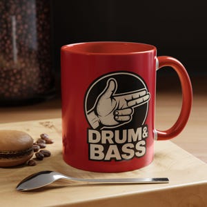 Puede incluir: Una taza de cerámica roja con un gráfico en blanco y negro de un gesto con la mano y las palabras "DRUM & BASS". La taza está sobre una tabla de madera con una cuchara y un macaron.