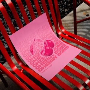 Puede incluir: Una impresión rosa con una ilustración de cerezas y las palabras "Cherry Bomb" en un patrón repetido. La impresión está sobre una silla de metal roja. También es visible el texto "Pastel Lunar".