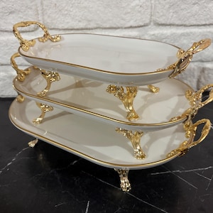 Puede incluir: Una bandeja de servir de tres niveles con un diseño ovalado blanco y adornos y asas doradas. La bandeja está sostenida por patas doradas ornamentadas. Las bandejas están apiladas una encima de la otra.