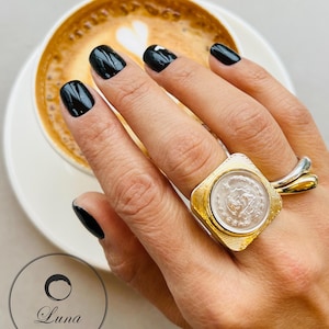 Pode incluir: Um anel dourado com um centro quadrado em forma de moeda e uma banda texturizada. O anel está numa mão com verniz de unhas preto, ao lado de uma chávena de café com um desenho em forma de coração na espuma. O logótipo Luna Accessories está no canto inferior esquerdo.