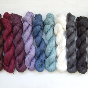 May include: Ten skeins of yarn arranged in a row, featuring a spectrum of colors including dark brown, deep red, plum, lavender, navy blue, teal, white, charcoal gray, black, and light beige, each with a twisted texture and fiber sheen.