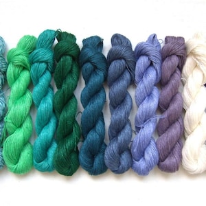 Puede incluir: Una colección de nueve madejas de hilo en una variedad de colores, incluyendo tonos de verde azulado, verde, azul, morado, blanco y negro. El hilo está fuertemente torcido y parece suave, ideal para proyectos de tejido o ganchillo.
