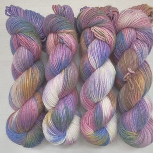 Pode incluir: Quatro novelos de fio tingido à mão em uma gama de cores com tons de roxo, rosa, azul, branco e marrom.