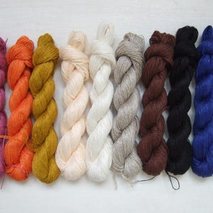 Puede incluir: Diez madejas de hilo retorcidas dispuestas en fila, con una vibrante paleta de colores que incluye magenta, naranja, amarillo mostaza, crema, blanco, gris claro, marrón oscuro, negro, azul real y verde azulado, con una textura suave y brillante.