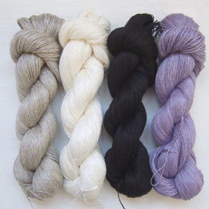 Könnte beinhalten: Vier Garnstränge in einer Reihe, jede in einer anderen Farbe. Von links nach rechts sind die Farben ein helles Graubraun, Off-White, Schwarz und ein gedämpftes Lila. Das Garn ist zu einer seilartigen Form verdreht.