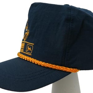 May include: A navy blue baseball cap featuring a gold braided cord across the base of the crown. The front panel displays an embroidered gold logo with the text "JOHN DEERE" and a leaping deer graphic, showcasing a structured design and visor.
