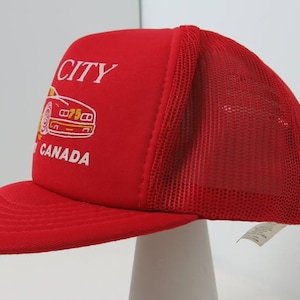 May include: A bright red trucker cap featuring a mesh back panel and a foam front panel. The front displays a white embroidered graphic of a car with the text CITY and CANADA printed in white lettering above and below the car illustration.
