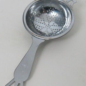 May include: A stainless steel tea infuser with a perforated bowl and a long handle. The handle has a decorative geometric design at the end and a small decorative element at the top of the bowl. The tea strainer is designed for steeping loose leaf tea.