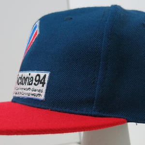 May include: A navy blue baseball cap with a red peak. The cap has a logo with red, white, and blue details and the text "Victoria 94 Commonwealth Games".