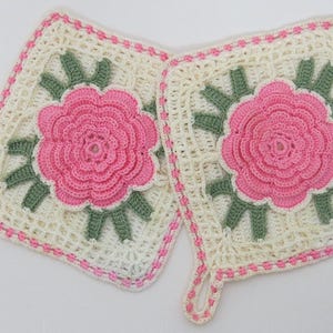 May include: Two handmade crochet potholders. Each square potholder features a pink rose in the centre, surrounded by green leaves. The potholders are cream-coloured with pink trim and a loop for hanging.