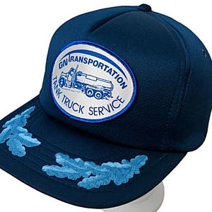 May include: A navy blue trucker cap with a white and blue patch displaying "GN Transportation Tank Truck Service." The brim has light blue leaf embroidery.