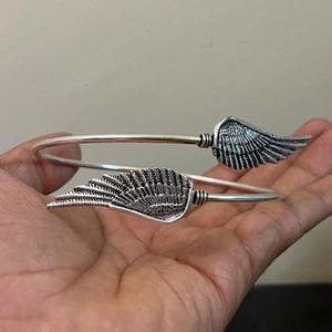 May include: A silver-toned cuff bracelet featuring two detailed wing designs. The wings are positioned on either side of the open bracelet, creating a symmetrical and elegant look. The bracelet is held in a hand, showcasing its design.