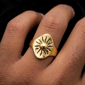 May include: A gold-colored ring featuring a sunburst design. The ring has a rounded rectangular face with a central raised sphere, surrounded by radiating lines. The ring is worn on a finger, showcasing its intricate details and warm metallic hue.