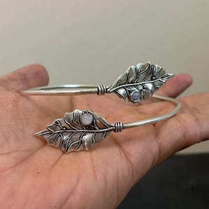 May include: Silver-toned cuff bracelet featuring leaf-shaped accents. Each leaf has intricate detailing and a small, round, light-colored stone. The bracelet is open, allowing for easy wear and adjustment. A nature-inspired jewelry piece.