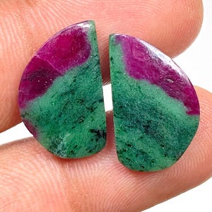 Puede incluir: Dos piedras preciosas pulidas, cada una con forma de media luna. La parte superior de cada piedra es de un color magenta intenso, que pasa a una base verde vibrante. Las piedras se sostienen entre los dedos.