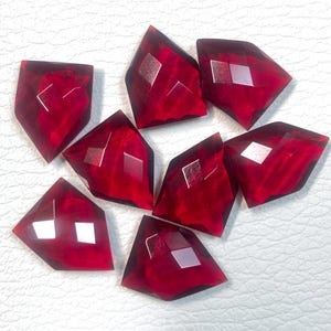 May include: Eight faceted, shield-shaped, deep red gemstones. Each gem has a polished, reflective surface with a diamond-shaped facet in the center. The gems are arranged on a white surface.