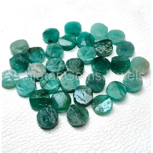 May include: A collection of small, circular, flat-cut emerald gemstones in various shades of teal and forest green. Each stone features a polished, reflective surface with natural inclusions and variations in color intensity and translucency.