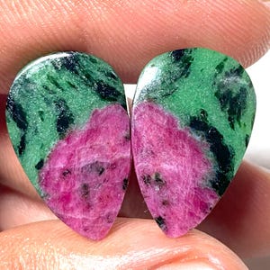 May include: Two teardrop-shaped gemstones. Each stone features a vibrant mix of colors: a vivid pink at the base, transitioning to a green upper section with black inclusions. The stones are polished and smooth, showcasing natural patterns.