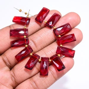 May include: A strand of faceted, rectangular, deep red gemstone beads. The beads are strung together with gold-colored spacers and wire, creating a necklace or bracelet component. The gemstones are held in a hand against a white background.