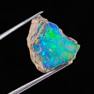 Puede incluir: Una gema de ópalo en bruto con vibrantes colores iridiscentes azules, verdes y amarillos. El ópalo está sujeto por unas pinzas plateadas sobre un fondo negro. La piedra tiene una textura rugosa y natural.